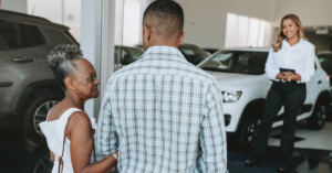 A man walking his mom into a car dealership with a sales rep smiling and ready to discuss vehicle loans.